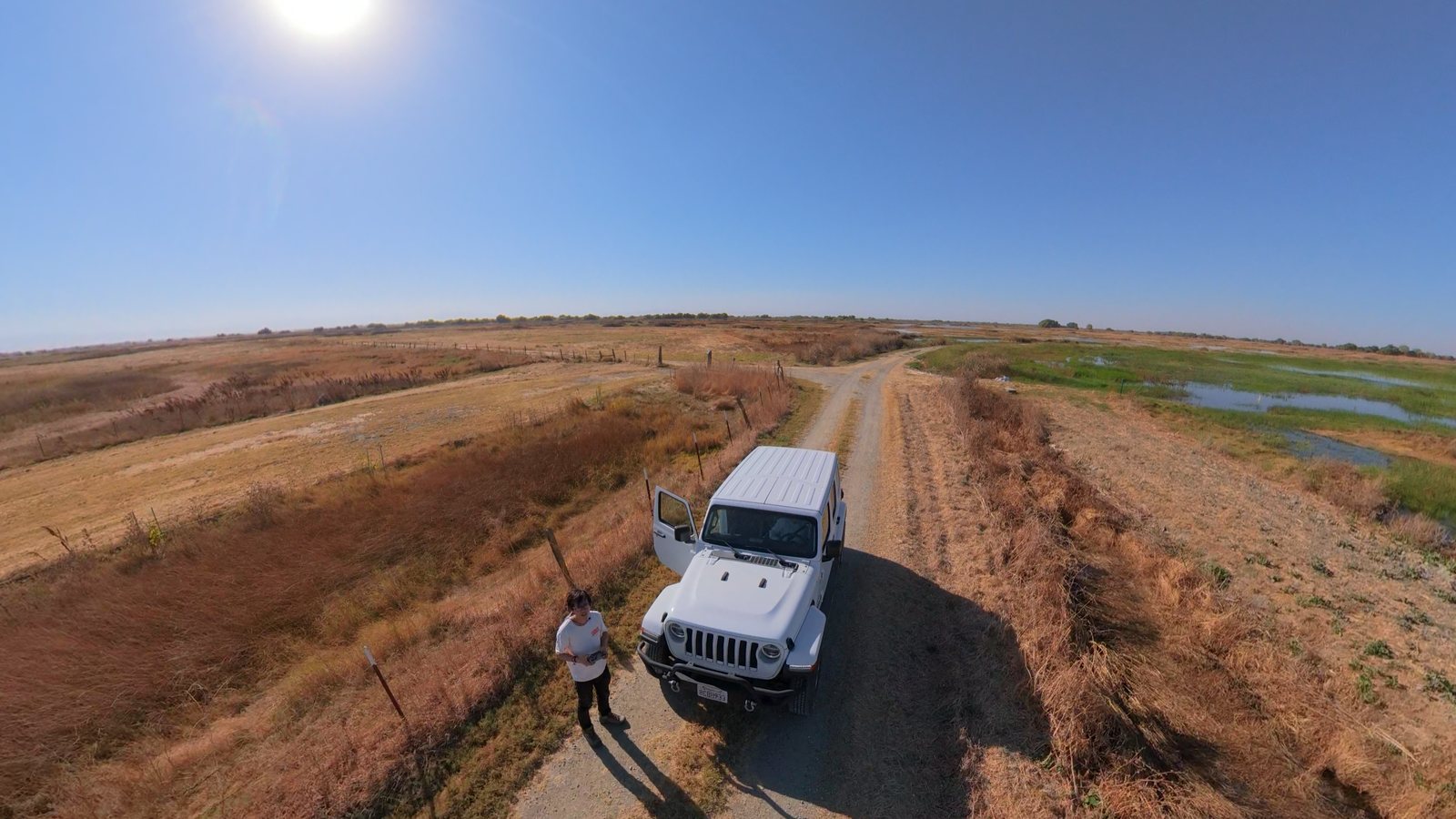 An Avata 360 drone shot reframed to 16:9: looking down at a white Jeep and its driver on a dirt track through golden marshland.